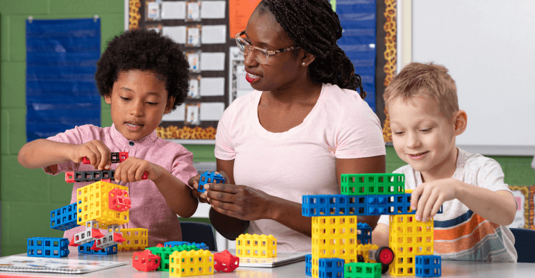 Black female teacher working with a young male Black student and a young male white student working with building blocks of many colors.