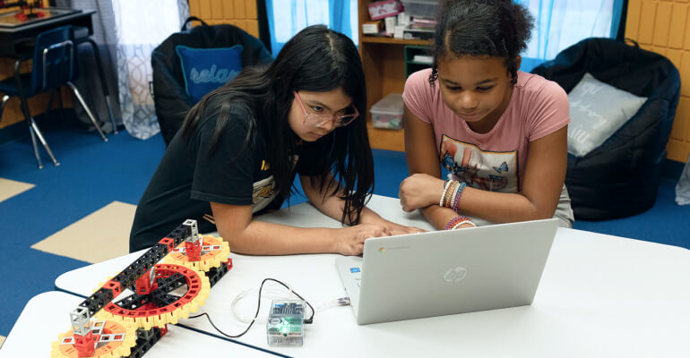 Two female middle school students work on a Kid Spark Education STEM lab. They assemble a robot that is connected to a laptop by a cable, while both girls work on the laptop.