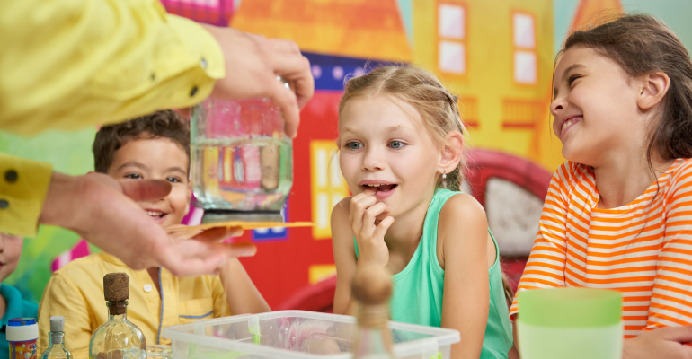 Two elementary school age female students and one male student sitting and observing an adult pair of hands that is holding a mason jar as part of a science experiment.