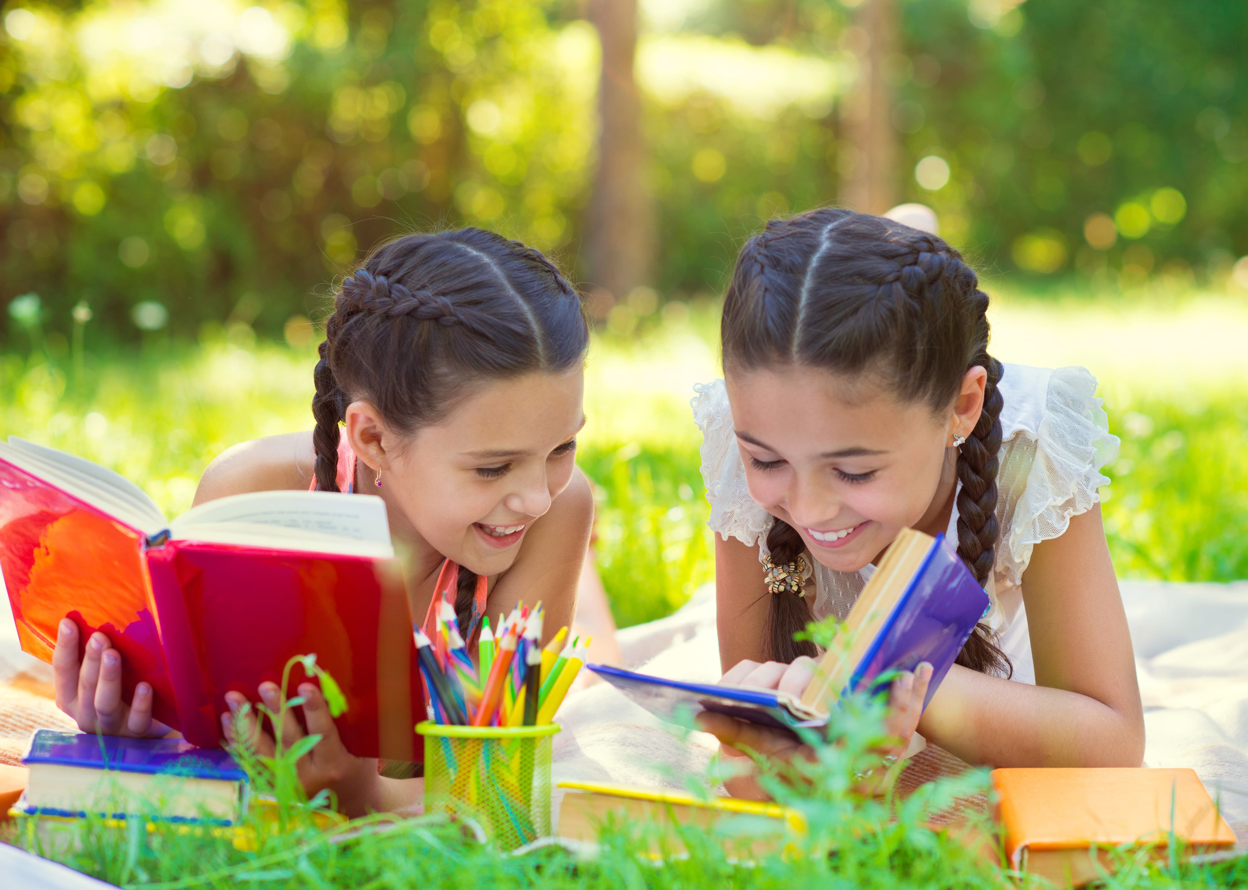 Two elementary age female students laying outside in a grassy field reading books.