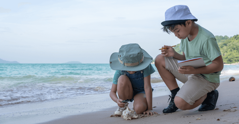 Two elementary aged students wearing hats, crouched on the beach. They are over a seashell that has washed ashore, with one student holding a magnifying glass to it and the other student holding a notebook and pencil.