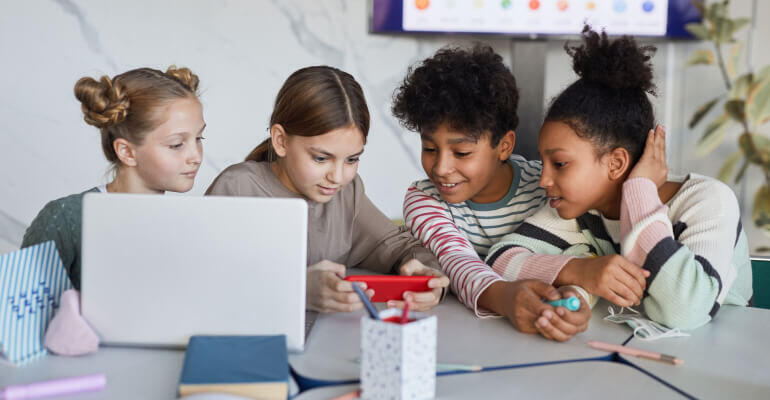 Group of four elementary age students sitting at some desks that are pushed together. They have a laptop open but look on as one of the girls holds a red smartphone in her hands and shows them something.
