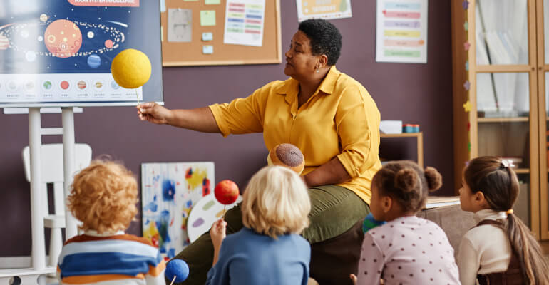 Teacher in a yellow shirt sits in front of a group of elementary age school children while she points at a chart of the solar system.