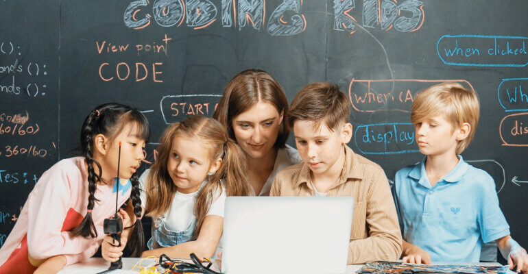 Four elementary school age students sit with a young female teacher in front of a chalkboard that has coding lesson notes written in chalk