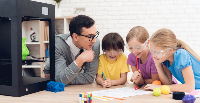 One male teacher with glasses sitting next two three young girl students all wearing safety goggles sitting around a worksheet and 3D pritner