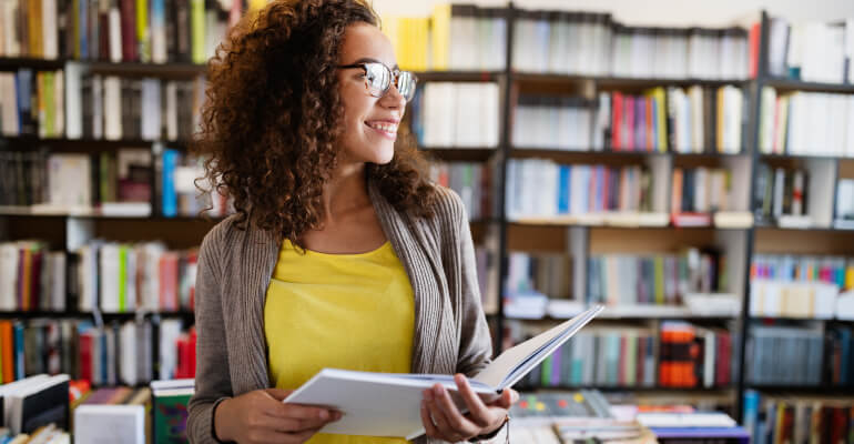 Young female educator standing in a library holding an open book. She is smiling and looking to the side.