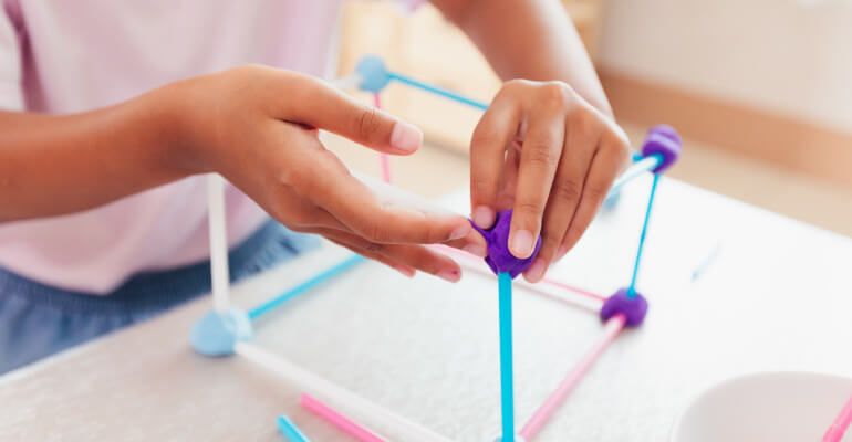 Close up of a student's hands building a cube with straws and purple clay