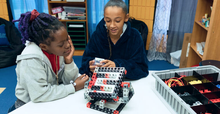 Two middle school aged female students sit at a desk in school. They are working on a STEM lab that features plastic blocks and wires that are being combined to work on robotics.