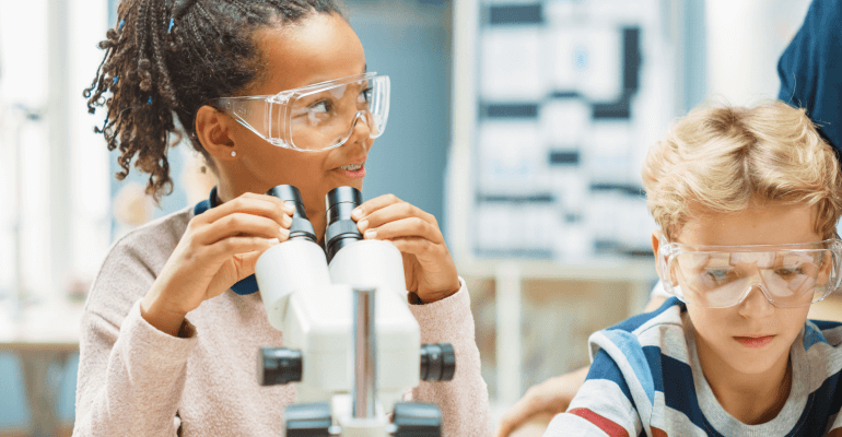 Young Black female student and young white male student sit with goggles on in front of microscopes.
