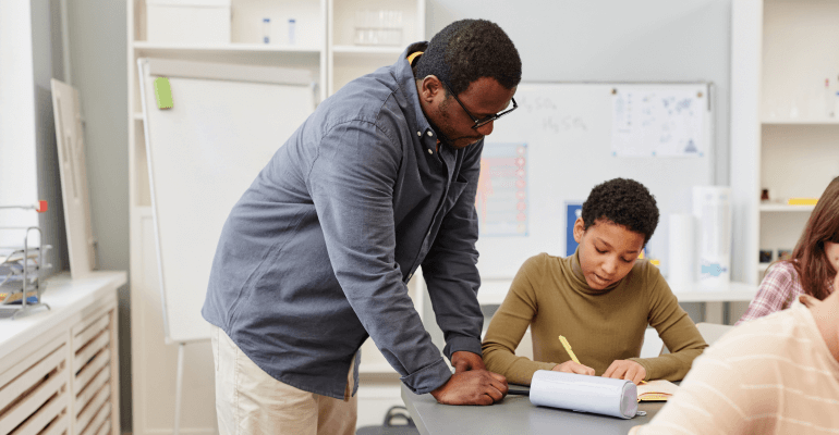 Black male teacher at a desk with a Black student as the student writes something down.