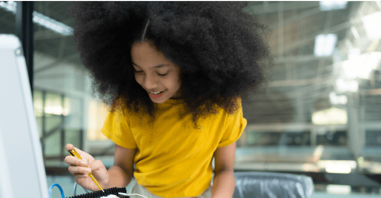 Young Black girl student in a bright yellow shirt smiles as she works on a robotic project.